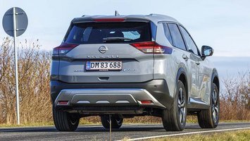 Nissan X-Trail set bagfra, mens den kører på en landevej med buske og et skilt i baggrunden
