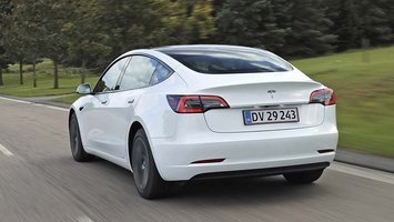 En hvis Tesla Model 3 set bagfra kører ad en landevej med træer i baggrunden.