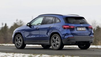 En blå Mercedes-Benz GLA 250 e set bagfra, mens den kører på en landevej med sne i forgrunden og træer i baggrunden.