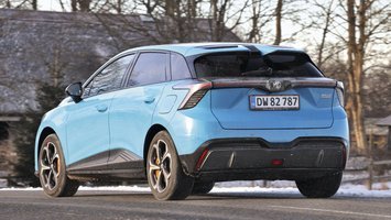 En lyseblå MG 4 XPower set bagfra, mens den kører på en vej med et hus i baggrunden