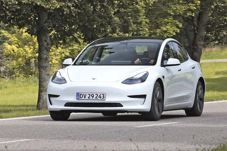 En hvid Tesla Model 3 set skråt forfra kører på en landevej med træer i baggrunden.
