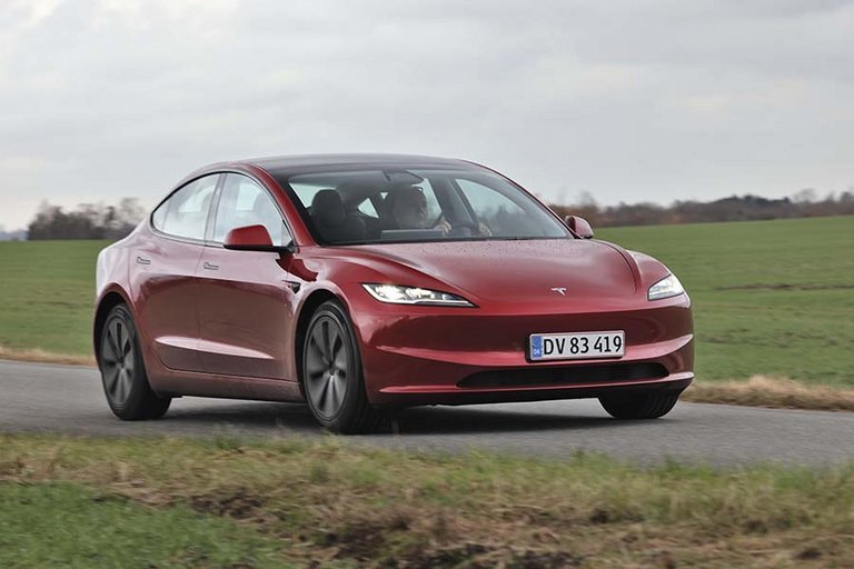 En rød Tesla Model 3 set forfra kører på landevej med græs i baggrunden