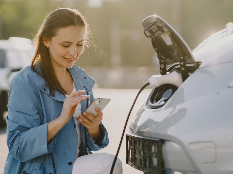 kvinde lader sin elbil ved ladestander og kigger på sin mobil. 