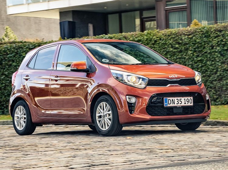 Orange Kia Picanto kører på brosten. I baggrunden en grøn hæk. 