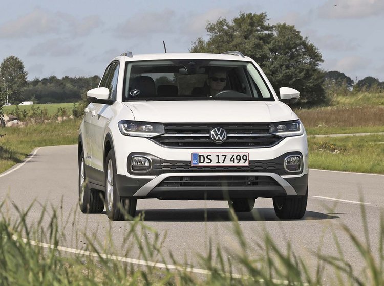 En hvid VW T-Cross mini-SUV kører på en landevej, der svinger. Græs i for- og baggrunden.