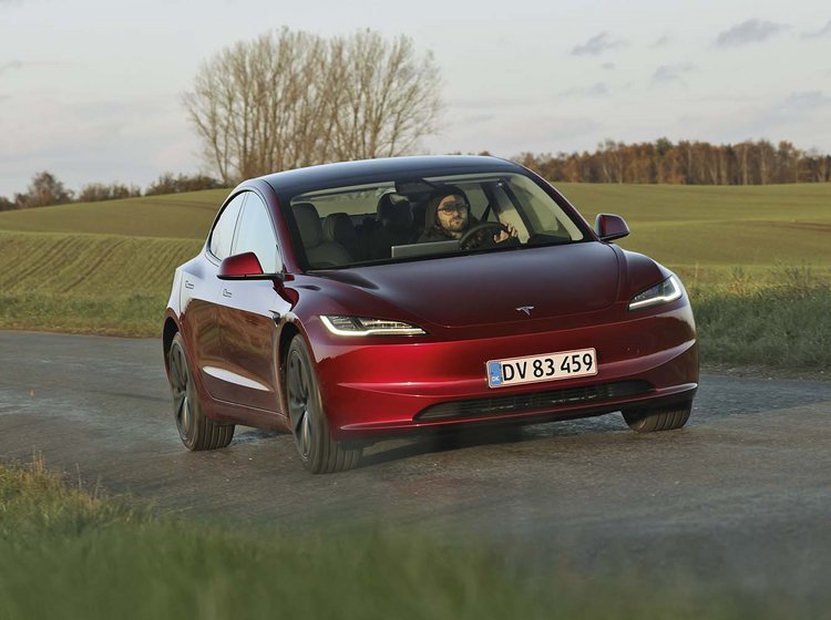 En rød Tesla Model 3 kører på en landevej med græs og et træ i baggrunden.