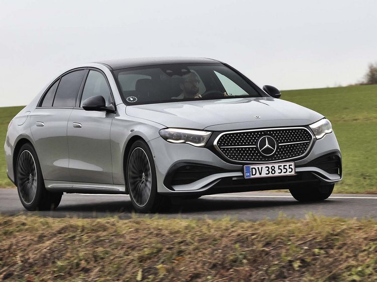 En sølvgrå Mercedes E-klasse set forfra, mens den kører på en vej med græs i baggrunden