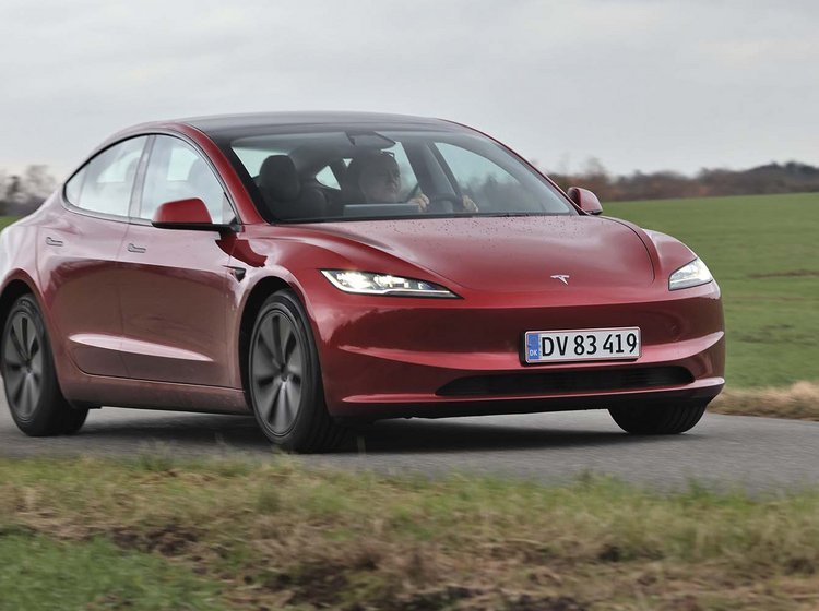 En rød Tesla Model 3 kører på en landevej med græs i baggrunden.