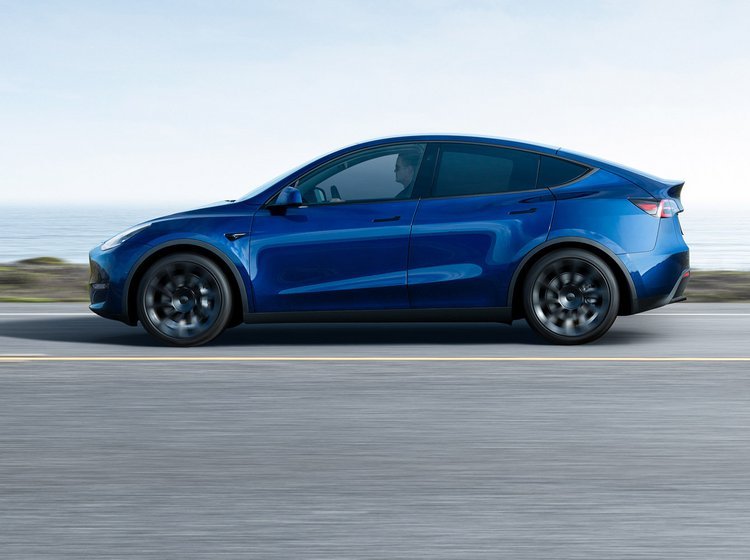 En blå Tesla Model Y fra siden. Kører på en landevej med vand i baggrunden. 
