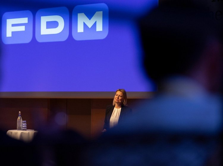 FDMs administrerende direktør, Stina Glavind, holder oplæg foran blå skærm med FDM-logo. Silhouetter af mennesker i forgrunden.