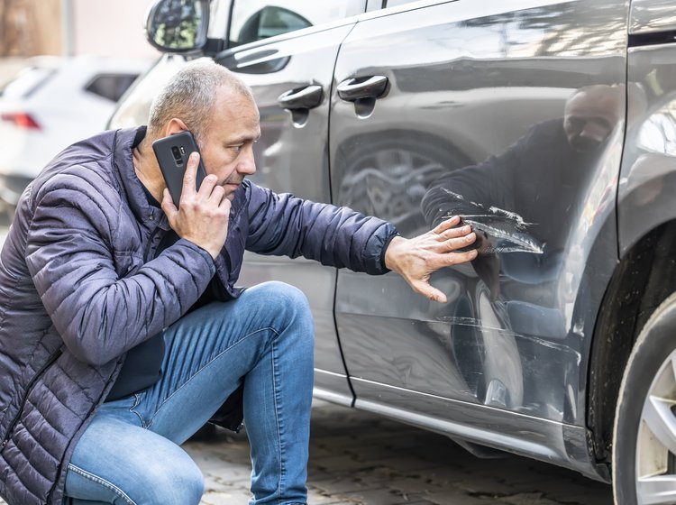 Mand taler i telefon, mens han kigger på en stor bule og ridse i sin bil.