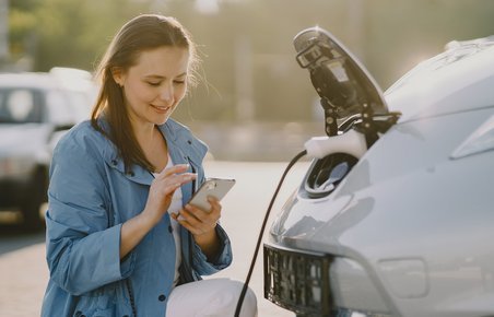 kvinde lader sin elbil ved ladestander og kigger på sin mobil. 