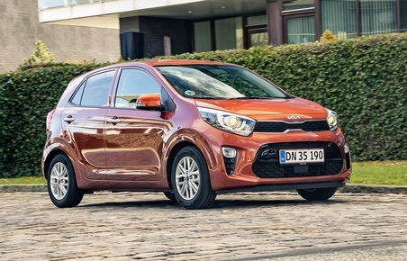 Orange Kia Picanto kører på brosten. I baggrunden en grøn hæk. 
