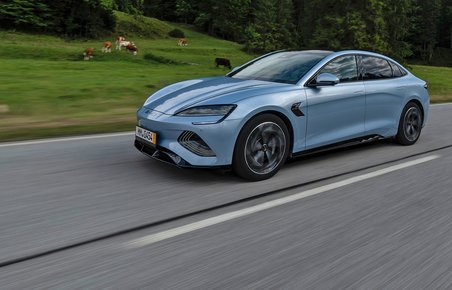 En lyseblå BYD Seal kører på en landevej med skove og bjerge i baggrunden