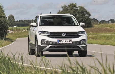 En hvid VW T-Cross mini-SUV kører på en landevej, der svinger. Græs i for- og baggrunden.