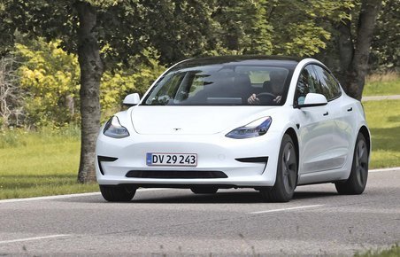 En hvis Tesla Model 3 kører på en landevej med græs og træer i baggrunden.