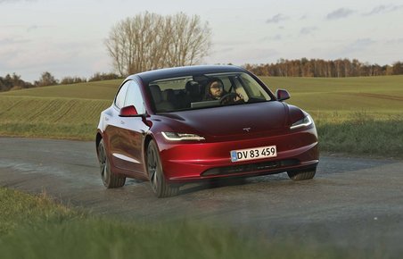 En rød Tesla Model 3 kører på en landevej med græs og et træ i baggrunden.