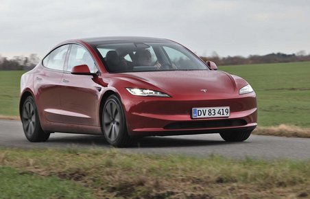 En rød Tesla Model 3 kører på en landevej med græs i baggrunden.
