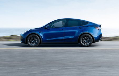 En blå Tesla Model Y fra siden. Kører på en landevej med vand i baggrunden. 