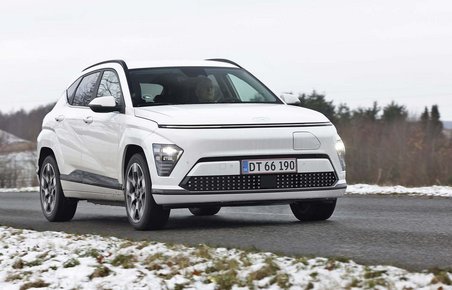 En hvid Hyundai Kona elbil kører på en landevej på en gråvejrsdag