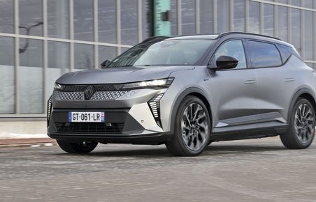 En mørkegrå Renault Scenic holder foran en bygning med mange vinduer