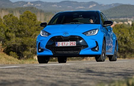 En blå Toyota Yaris kører på en vej med træer og bjerge i baggrunden