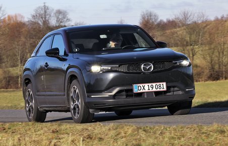 En sort Mazda MX-30 R-EV kører på en landevej med træer og græs i baggruden