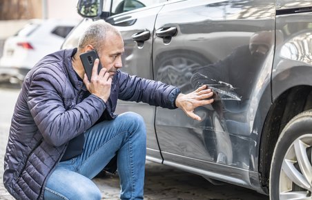 Mand taler i telefon, mens han kigger på en stor bule og ridse i sin bil.